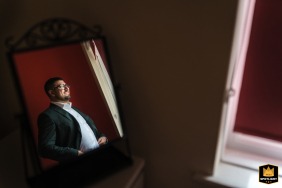 A wedding photographer captures a serene scene at a London hotel. The image, framed by a mirror, shows the groom getting ready by a window, bathed in soft, natural light.
